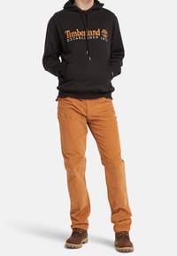 Persona che indossa un hoodie nero Timberland, pantaloni in velluto a coste arancio-marrone e stivali marroni, in piedi con le mani nella tasca dell'hoodie.