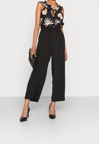 Pantaloni larghi neri abbinati a un top senza maniche con motivo floreale, dotato di scollatura arricciata e dettaglio a chiave, accompagnato da una pochette nera.