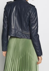 Giubbotto di pelle navy con un design corto, caratterizzato da spalline e una cerniera laterale, abbinato a una gonna verde plissettata.