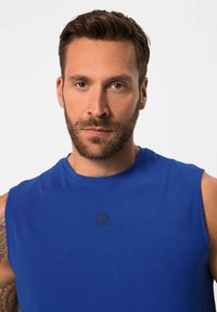 Blauw mouwloos shirt van gladde, zachte stof met een klein zwart logo op de borst gedrukt. Eenvoudig ontwerp met een ronde halslijn.