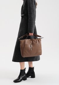 Brązowa skórzana torebka o fakturowanym wykończeniu, z dwoma uchwytami u góry i zapięciem na zatrzask, trzymana przez osobę ubraną w węgorzowaty płaszcz i czarne botki.