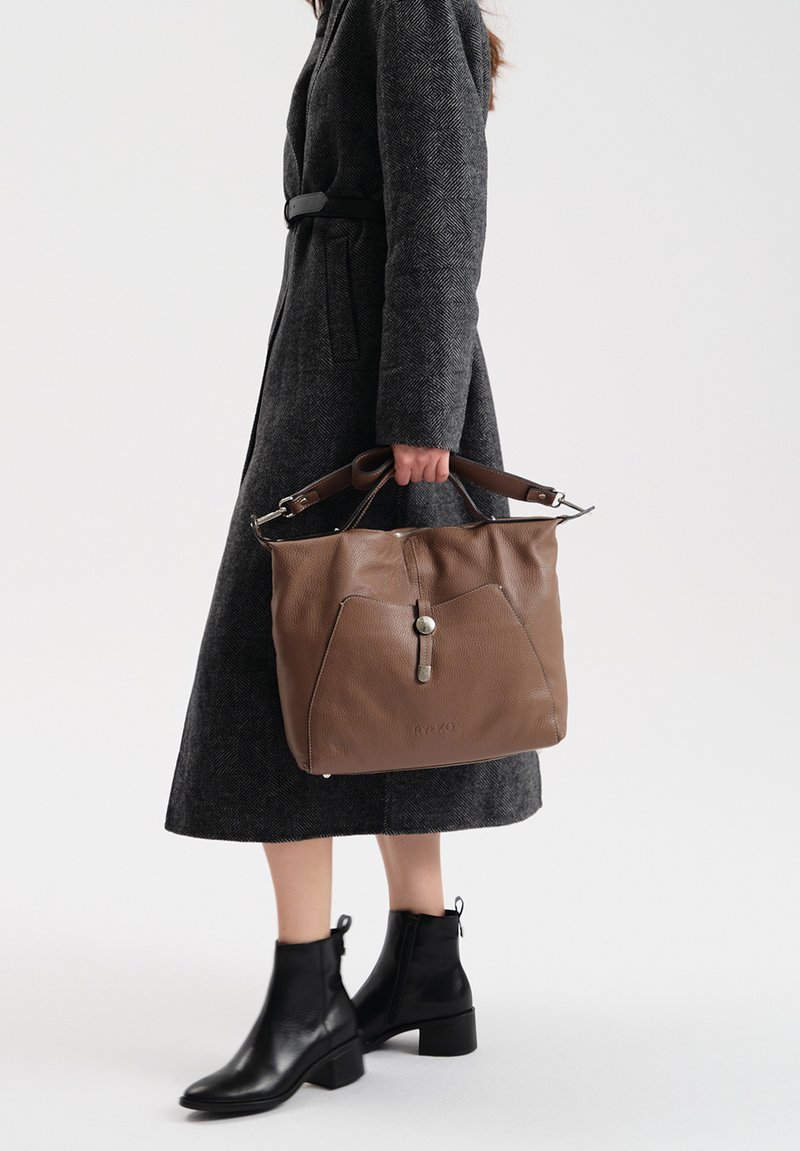 Brązowa skórzana torebka o fakturowanym wykończeniu, z dwoma uchwytami u góry i zapięciem na zatrzask, trzymana przez osobę ubraną w węgorzowaty płaszcz i czarne botki.