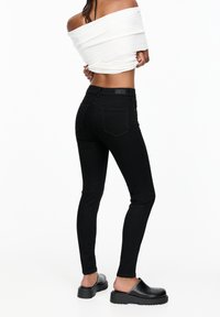 Osoba oblečená v černých skinny jeans, černých slip-on platform botách a bílém krátkém topu s odhalenými rameny na bílém pozadí.