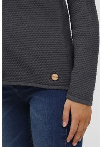 Suéter gris de punto con tejido texturizado, logo de madera redondo en el dobladillo y puños acanalados, combinado con jeans de mezclilla azul.