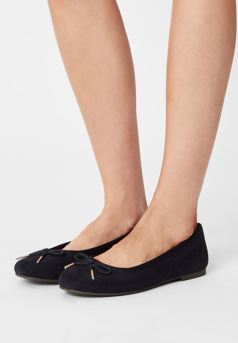 Paire de ballerines noires avec des nœuds décoratifs portées pieds nus sur un fond blanc uni.