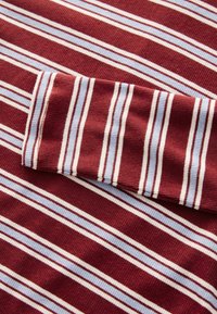 Gros plan sur un tissu côtelé à rayures horizontales bordeaux, blanc et bleu clair, avec un détail de manche pliée.