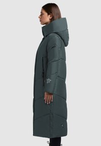 Tamnozeleni kaput s punjenjem i kapuljačom, kviltani dizajn, bočni džep s patentnim zatvaračem i detalji na kopčama. Dugačak i mekan na dodir.