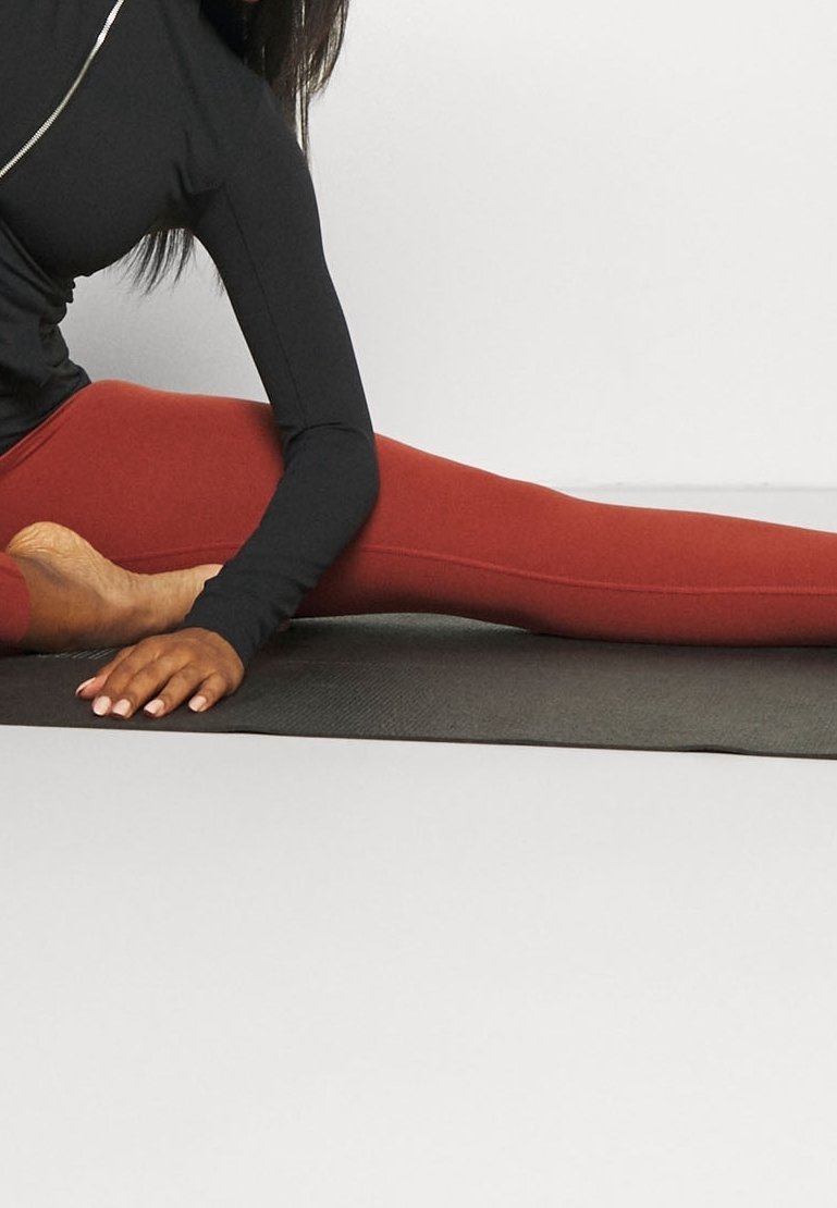 Sort langærmet top med lynlåsdetalje, parret med røde leggings. Emnet strækker sig på en tekstureret sort måtte. Fokus på design og farver inden for træningsbeklædning.