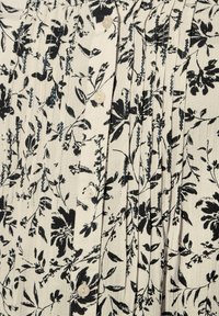 Blusa con stampa floreale su fondo crema, caratterizzata da fiori neri. Pieghe arricciate e quattro bottoni tondi creano texture e dettagli.