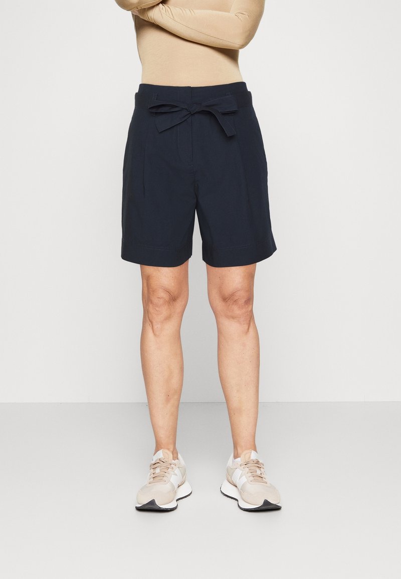 Shorts marine avec ceinture à nouer, dotés de plis sur le devant et d'une silhouette structurée. Associés à des chaussures de sport de couleur claire.