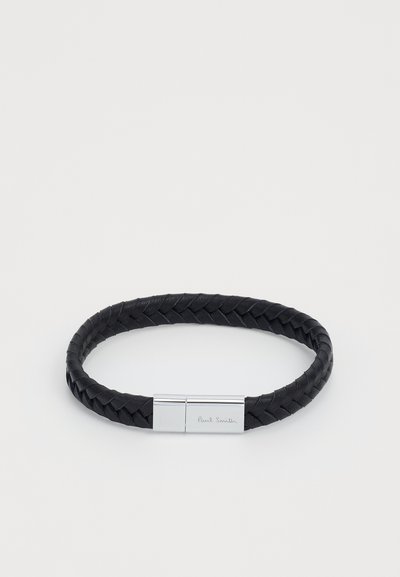 Sort flet læderarmbånd med en sølvfarvet lås, der har det indgraverede navn "Paul Smith." Glat tekstur og cirkulært design.