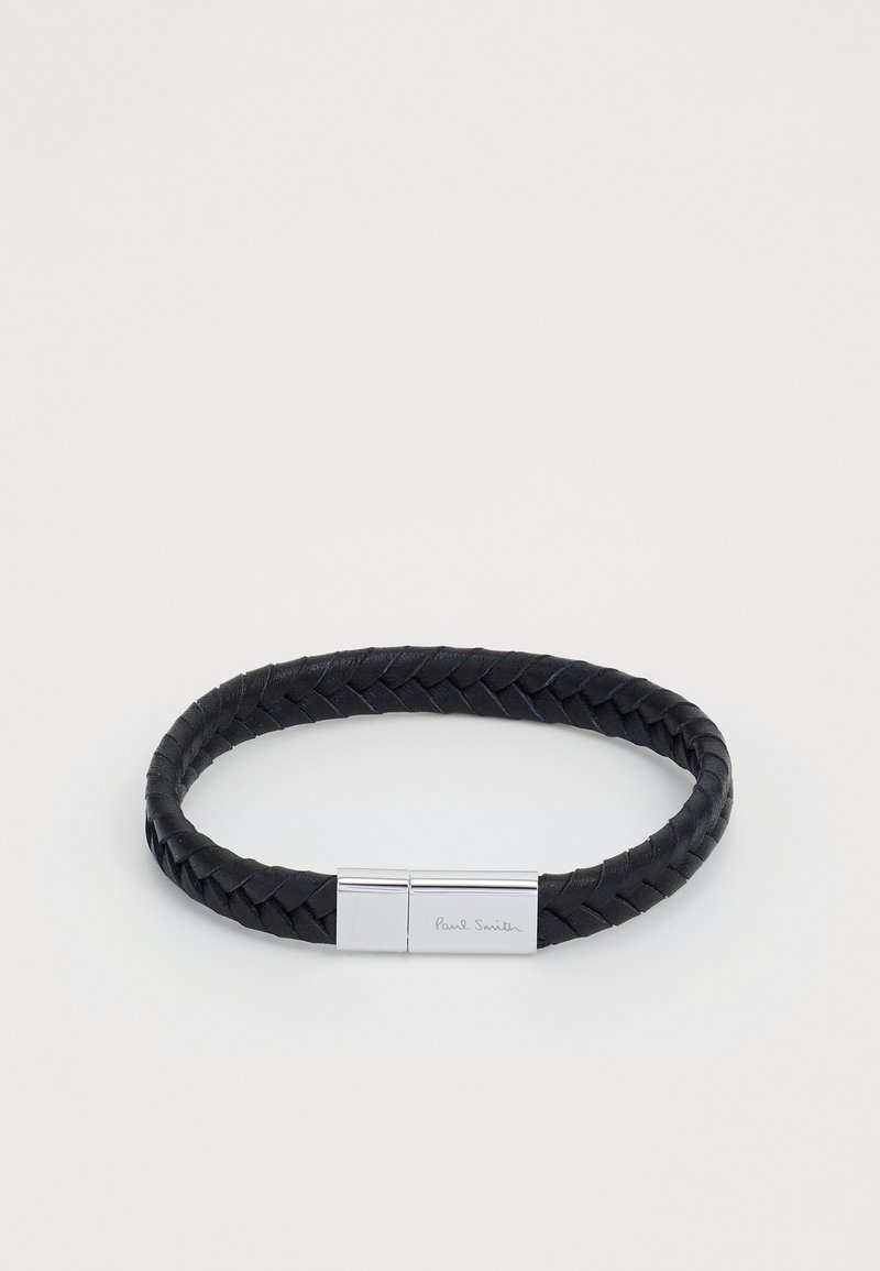 Melna pītā ādas aproce ar sudraba tonī izgatavotu aizdari, uz kuras ir iegravēts vārds "Paul Smith". Gluda virsma un apaļa forma.