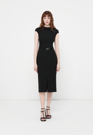 Young woman standing facing forward wearing a fitted black midi dress with cap sleeves, a waist belt, and black T-strap heels.