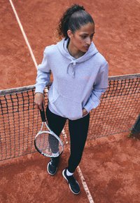 Felpa grigia con tasca frontale e cordoncino, leggins neri e scarpe nere, con una racchetta da tennis in mano su un campo in terra battuta.