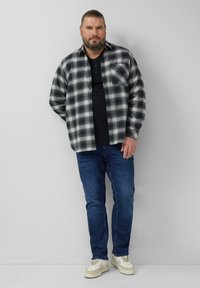 Brodaty mężczyzna z długimi włosami związanymi z tyłu, ubrany w czarno-białą koszulę w kratę na czarnej koszulce, niebieskie dżinsy i białe trampki, stoi opierając się o ścianę.