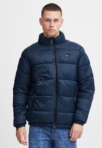 Mørkeblå pufferjakke med høj krave, quiltet design, frontlynlås og sidelommer. Lavet af glat, let materiale.