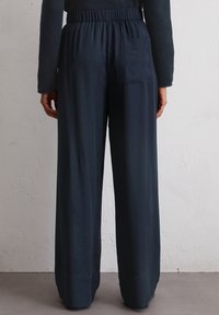 Marineblauwe wijde broek met een elastische tailleband, een gladde textuur en een achterzakdetail. De stof valt soepel om de enkels.