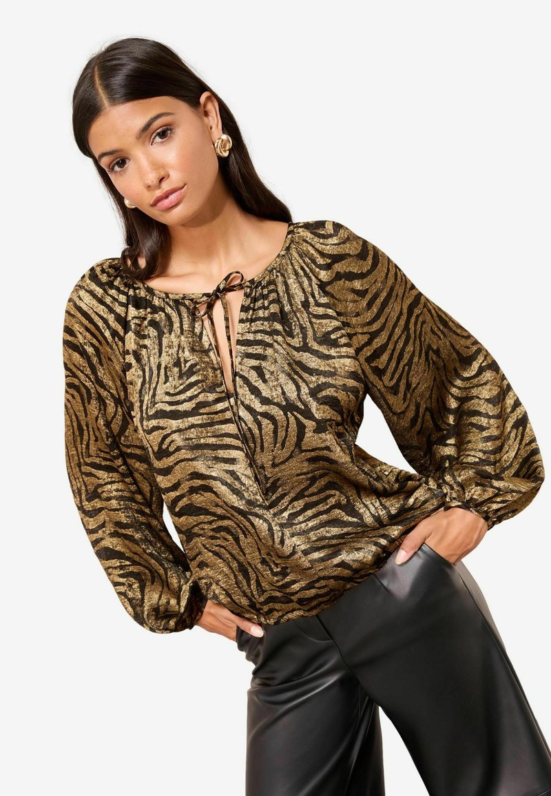 Metallisch goldene und schwarze Bluse im Zebra-Design mit einem Krawatten-Ausschnitt und weiten Ärmeln, kombiniert mit schwarzen Lederhosen.