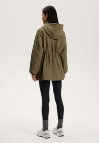 Vihreä vuorattu takki hupulla, jossa on kiristysnyöri vyötäröllä ja pitkät hihat. Päällä mustat leggingsit ja harmaat sukat.