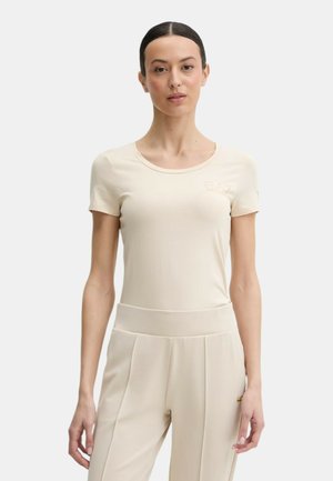 Donna con capelli scuri raccolti, che indossa una maglietta beige a maniche corte aderente e pantaloni beige a vita alta, in piedi davanti a uno sfondo semplice.