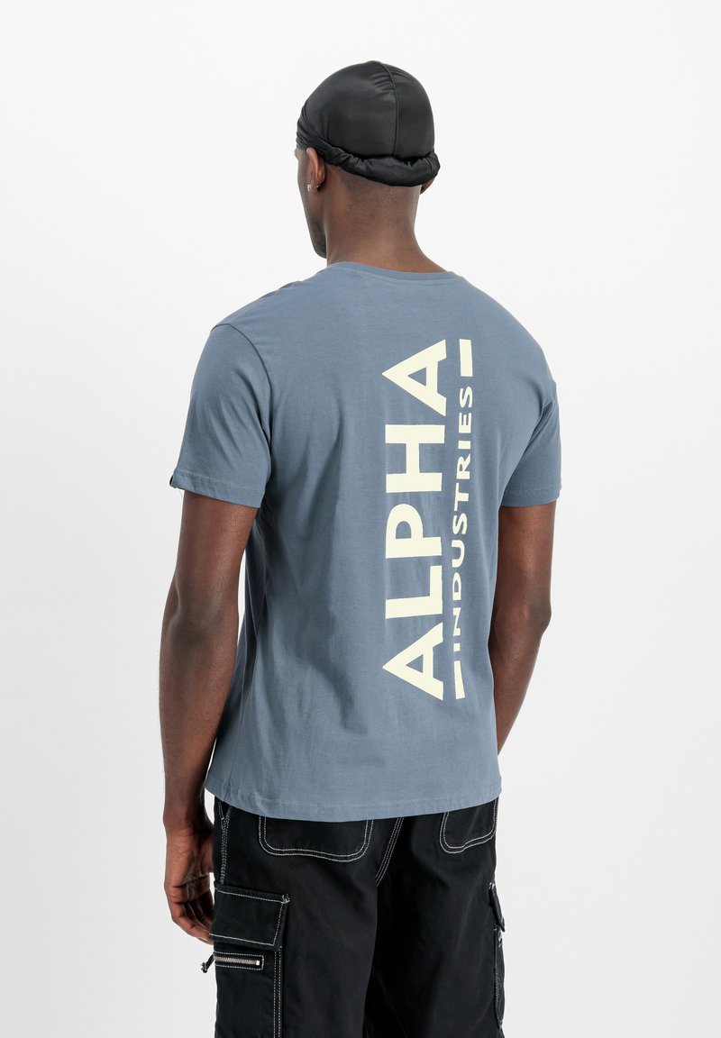Kék pamut póló nagy krém színű grafikai szöveggel, amely a hátán található: "ALPHA INDUSTRIES", kerek nyakkivágással és rövid ujjakkal.