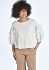 Vit kortärmad topp med puffärmar, tillverkad i mjuk bomull, med avslappnad passform och rund halsringning. Bärs med beige byxor.