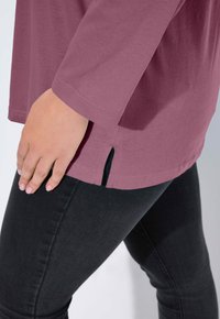 Mauve shirt met lange mouwen en een zijsplit, gedragen over donkere skinny jeans. De stof is glad met een licht elastische textuur.