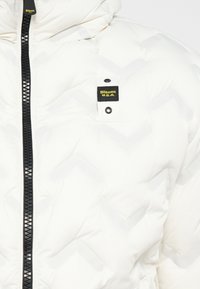 Witte gewatteerde pufferjack met een zwarte rits en hardware. Beschikt over een borstzak met een geel en zwart label. Geometrisch patroon zichtbaar.
