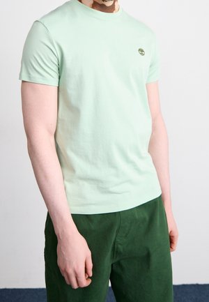 Mand iført lysegrøn kortærmet t-shirt med lille logo på brystet og mørkegrønne bukser, stående foran en ensfarvet lys baggrund.