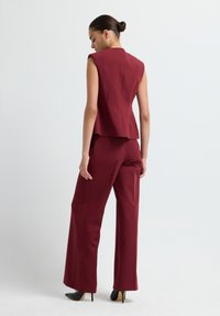 Top sans manches taillé bordeaux avec un design structuré, associé à un pantalon large, fabriqué à partir d'un tissu lisse. Des escarpins noirs à bout pointu accentuent la tenue.
