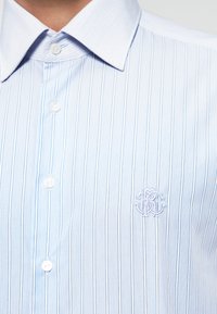 Světle modrá pruhovaná košile s klasickým límcem, zapínáním na knoflíky a malým vyšitým logem na hrudi. Vyrobena z lehké látky.