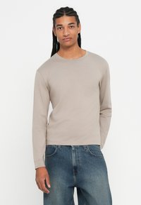 Man met gevlochten haar, gekleed in een beige langslurfshirt en blauwe jeans, staat met één hand in zijn zak tegen een witte achtergrond.