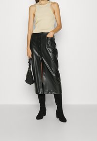 Musta keinonahkainen midi-hame, jossa on edessä halkio, sivutaskut ja nappiyksityiskohtia, yhdistettynä beigeen neuletoppiin ja mustiin saappaisiin.