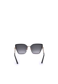 Lunettes de soleil angulaires à monture noire avec verres à dégradé foncé et branches fines dorées posées sur une surface blanche.