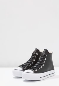 Sorte skinn høye sneakers med hvite gummisåler og tåhette, med svarte snører og sølvfargede hull, glatt tekstur.