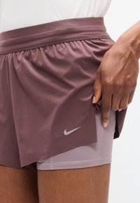 Σορτς Nike σε μοβ απόχρωση, με στρωματοποιημένο σχέδιο. Η εξωτερική στρώση είναι ελαφριά με ελαστική μέση και ανοίγματα στα πλάγια· η εσωτερική στρώση είναι σε ανοιχτό μωβ χρώμα.