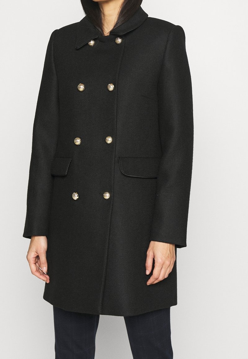Abrigo negro de doble botonadura con una textura tejida, que presenta botones en tono dorado, cuello ancho y dos bolsillos frontales.