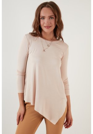 Top a maniche lunghe color pesca chiaro con orlo asimmetrico, scollatura rotonda e texture morbida, abbinato a pantaloni beige e collane a strati.