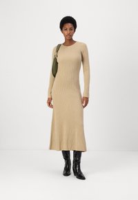 Abito a maglia beige a coste con maniche lunghe, busto aderente e orlo svasato; abbinato a una borsa a spalla verde e stivali neri sopra il ginocchio.