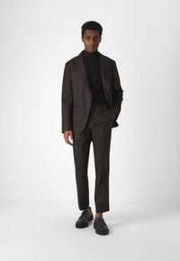Brun skreddersydd dress med en enknapps blazer over en svart turtleneck. Kombinert med svarte loafers. Slim fit bukser med glatt tekstur.