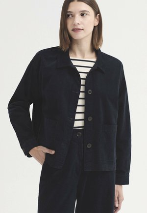 Femme portant une veste noire boutonnée sur une chemise rayée blanche et noire, une main dans la poche, regardant légèrement sur le côté.