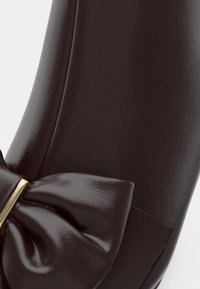 Détail de chaussure en cuir marron foncé avec un grand nœud assorti et un accent métallique doré à l'avant.