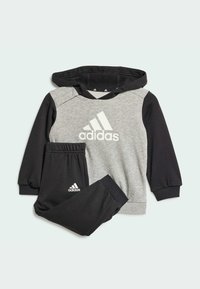 Felpa Adidas grigia e nera con logo bianco, dotata di cappuccio e polsini a coste, abbinata a pantaloni della tuta neri.