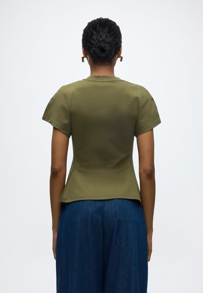 Γυναίκα με την πλάτη στην κάμερα, φοράει ελιάς πράσινη κοντομάνικη μπλούζα και μπλε παντελόνι φαρδύ, μαλλιά σε κότσο, με χρυσά γεωμετρικά σκουλαρίκια.