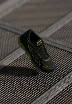 Zwarte en neon groene ASICS hardloopschoen hangend boven een gestructureerd metalen roosteroppervlak.