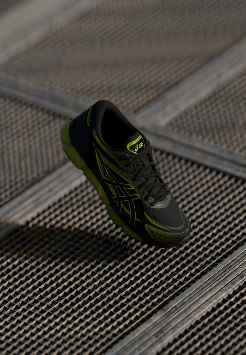 Chaussure de course ASICS noire et vert néon suspendue au-dessus d'une surface en grille métallique texturée.