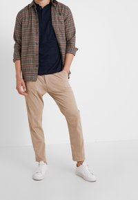 Koszula flanelowa w kratę w ziemistych odcieniach, ciemne polo, beżowe spodnie oraz białe trampki. Koszula zapinana na guziki z podwiniętymi rękawami.