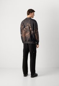 Mand med sorte cargo-bukser og en mørk sweatshirt med tryk af balletdansere på ryggen, stående mod en enkel hvid baggrund.