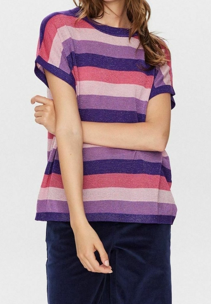 Femme portant un haut à manches courtes, ample et rayé dans des tons violet, rose et rouge, associé à un pantalon bleu foncé, bras croisés sur la poitrine.