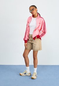 Różowa satynowa kurtka z czerwonymi akcentami i trzema paskami, biała koszulka, beżowe szorty oraz beżowe sneakersy z ciemnoniebieskimi akcentami. Przezroczyste okulary przeciwsłoneczne.